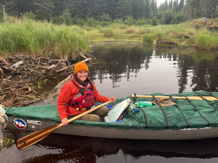 woman holds her Bending Branches canoe paddle sitting in a loaded canoe on a narrow river